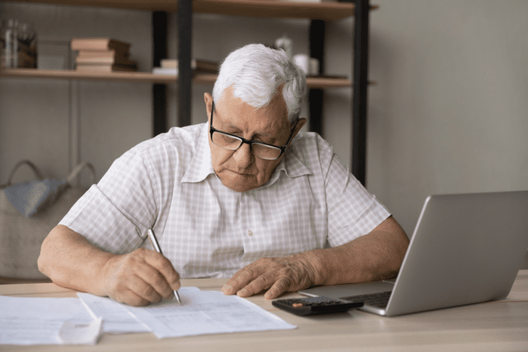 An old man with white hair sits at a desk with a laptop, calculator and paper