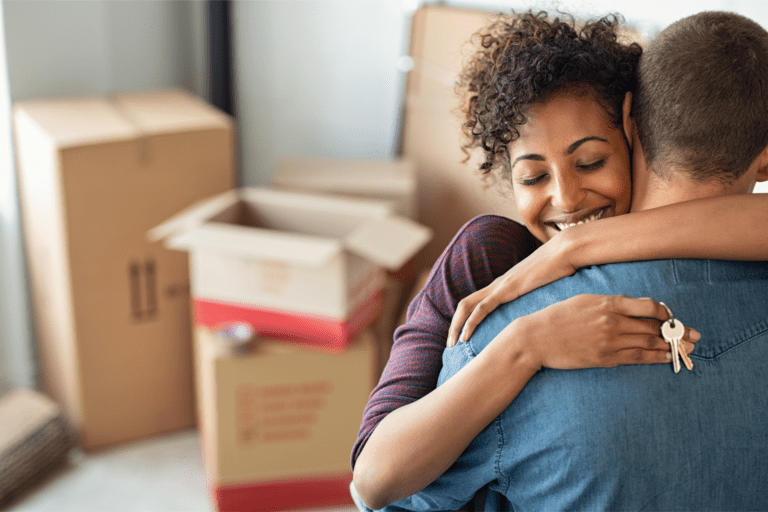 Black woman with curly black hair embraces a short haired black man in a blue tee shirt. She is holding a set of keys and there are brown moving boxes in the background.